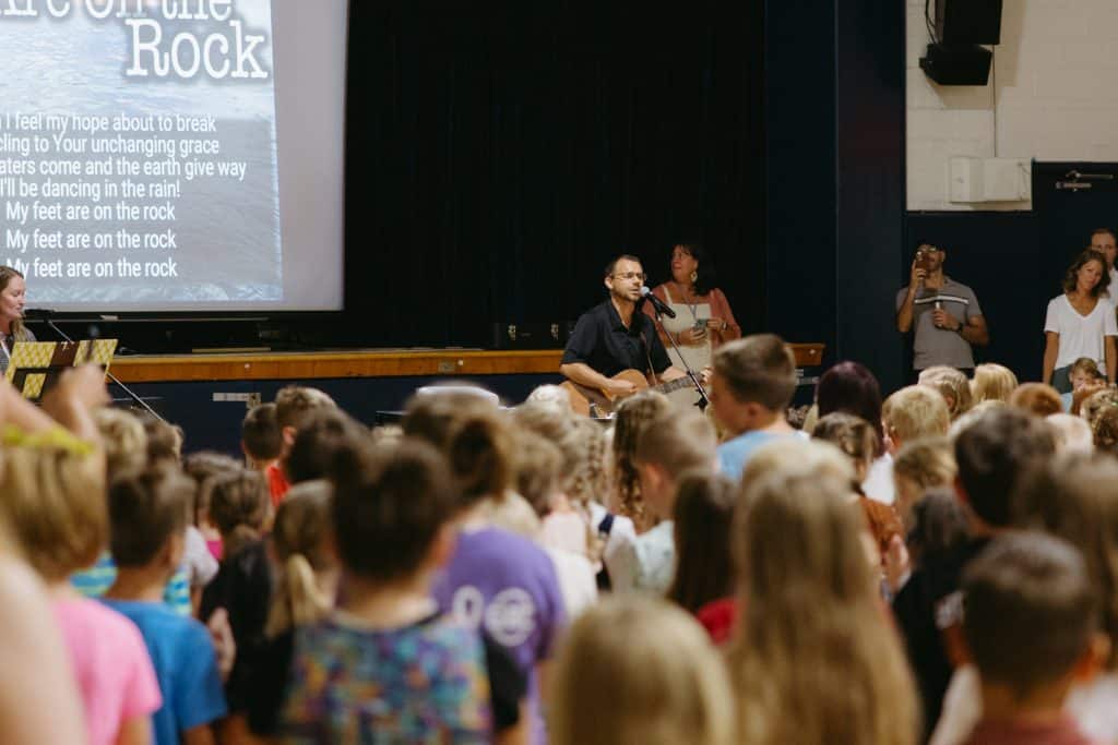 A staff member playing the guitar acoustically as they sing along to music projected onto a screen in the gym with kids attending