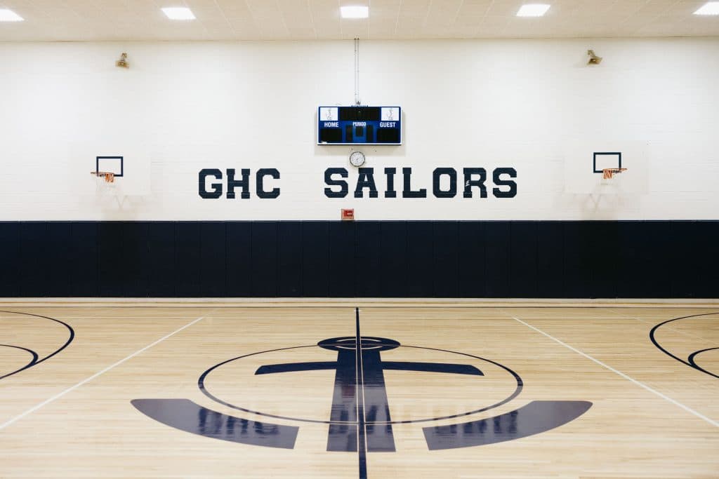 Empty gymnasium where you can see GHC Sailors on the wall with the shot clock and in the center of the court, the logo of a cross os visible