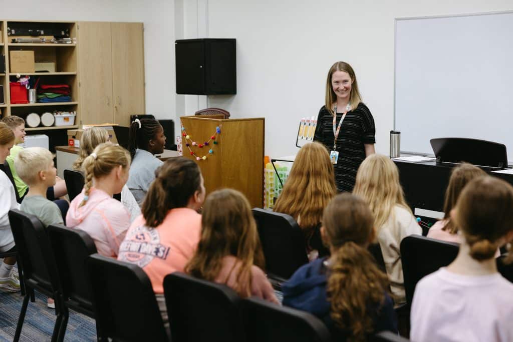 A teacher stands in front of her classroom as the kids sit and listen to her talk